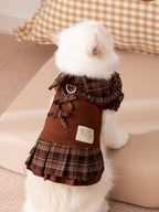 Back view of a white cat in a brown plaid pet dress with decorative bows and a D-ring leash attachment.