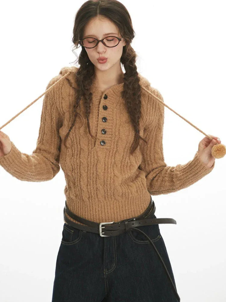 Close-up of a woman wearing a brown hooded cable knit sweater, holding drawstring with pom-pom
