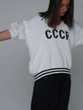 Back view of a woman wearing a white knit sweater with CCCP graphic print and striped collar detail.