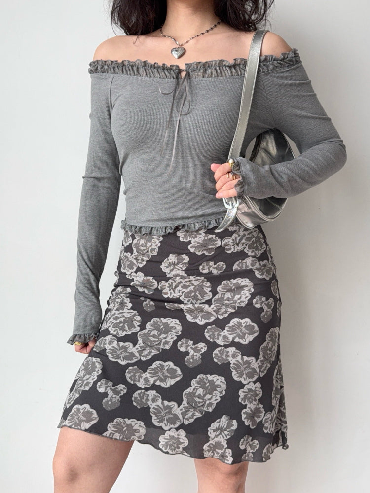 woman wearing a grey off-the-shoulder ribbed knit top with lace trim and a tie closure at the neckline, paired with a floral print skirt