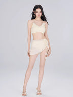 Woman wearing a cream-colored bikini top, wrap mini skirt, and sheer cover-up on a plain background.