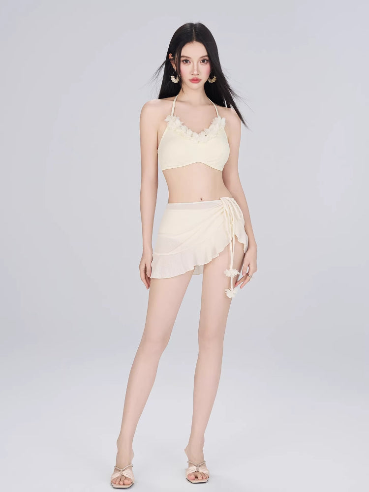 Woman wearing a cream-colored bikini top, wrap mini skirt, and sheer cover-up on a plain background.