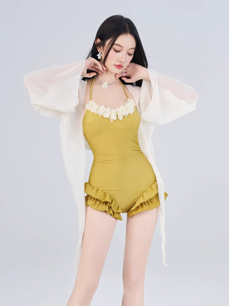 Woman posing in a mustard yellow one-piece swimsuit with floral trim and ruffled shorts, adjusting her necklace.