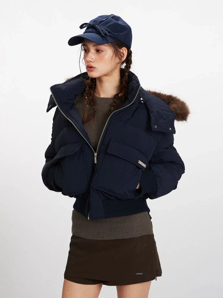 Front view of a woman wearing a navy blue cropped puffer jacket with a fur-trimmed hood, dark brown skirt, and brown boots.