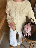 Close-up of a woman wearing a cream cable knit sweater and white pants, holding a coffee drink and a pink scarf in a brown handbag.