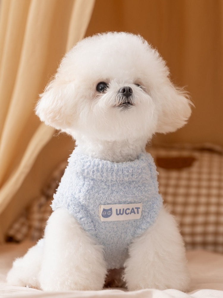 White bichon frise dog wearing a light blue fluffy dog sweater with a white "Wcat" logo patch on the chest.