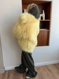 Woman wearing a yellow shaggy faux fur coat from the back, paired with dark green pants and a dark green hood.
