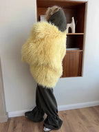 Woman wearing a yellow shaggy faux fur coat from the back, paired with dark green pants and a dark green hood.