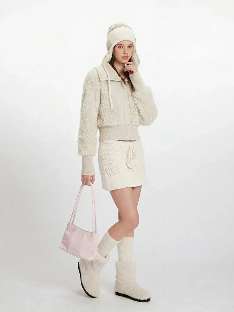 Full body shot of a woman in a cream fuzzy cropped jacket, white knit mini skirt, and pink handbag