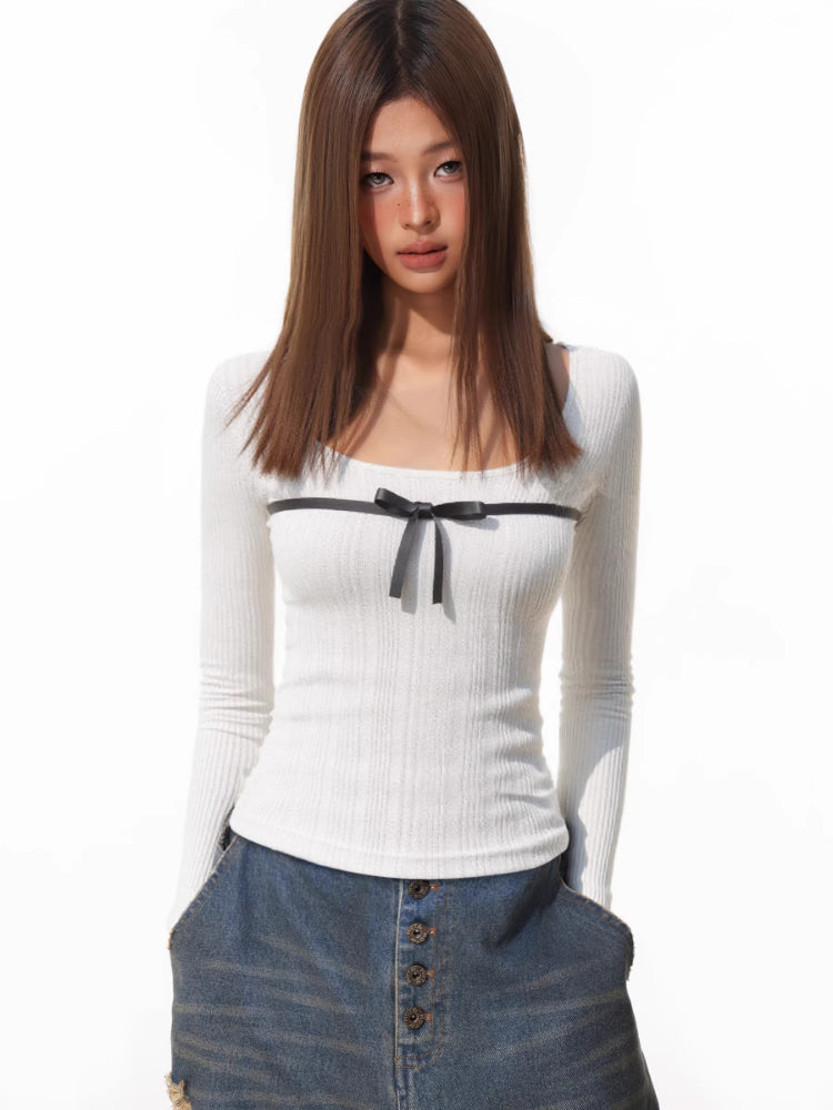 Woman wearing a white ribbed long-sleeve top with a black bow detail on the chest, paired with distressed denim shorts.