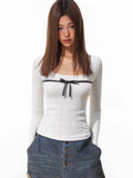Woman wearing a white ribbed long-sleeve top with a black bow detail on the chest, paired with distressed denim shorts.
