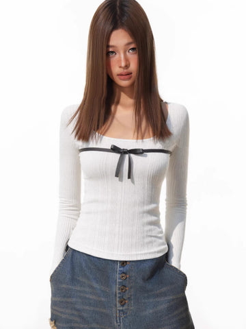 Woman wearing a white ribbed long-sleeve top with a black bow detail on the chest, paired with distressed denim shorts.