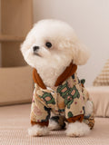 Small white dog wearing a tan patterned hooded dog jacket with brown trim.