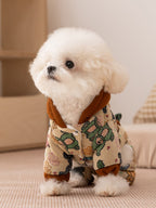 Small white dog wearing a tan patterned hooded dog jacket with brown trim.