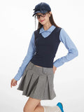 Woman posing in a blue striped collared shirt, navy V-neck vest, and grey pleated skirt, with glasses and a navy baseball cap.