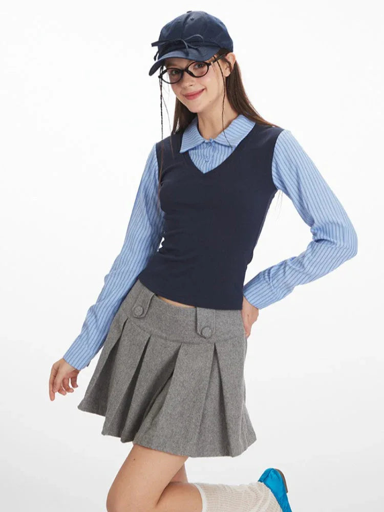 Woman posing in a blue striped collared shirt, navy V-neck vest, and grey pleated skirt, with glasses and a navy baseball cap.