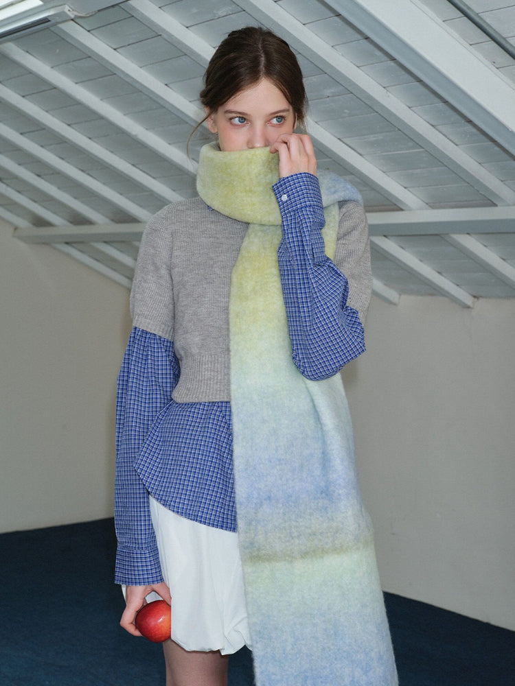 Woman wearing a long, oversized scarf with an ombre gradient of yellow, blue, and green, draped around her neck over a gray sweater and blue plaid shirt.