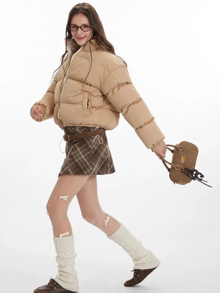 Woman in motion wearing a beige cropped puffer jacket, plaid skirt, and leg warmers.
