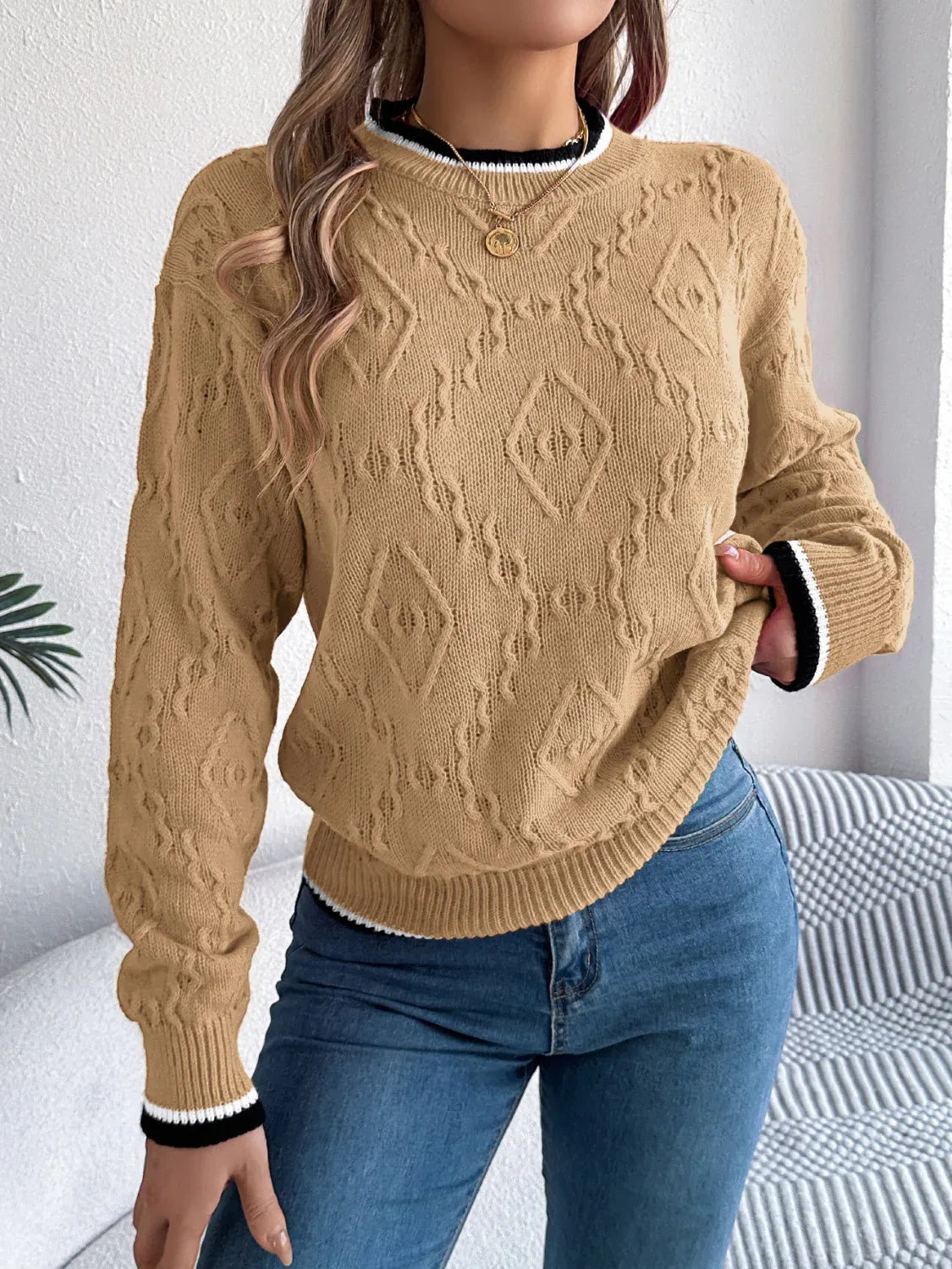 Woman wearing a tan cable knit sweater with a geometric pattern and contrast trim on the neckline and cuffs. Main image