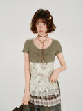 Woman wearing a green ribbed knit short sleeve top with a white and brown patterned corset-style overlay and lace trim.