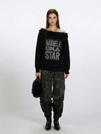 Woman wearing a black off-the-shoulder knit sweater with faux fur trim and a "NARIELE ON A STAR" graphic print, paired with camouflage cargo pants and black boots.