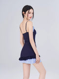Rear view of woman in navy one-piece swimsuit with blue gingham skirt overlay and white sheer cardigan