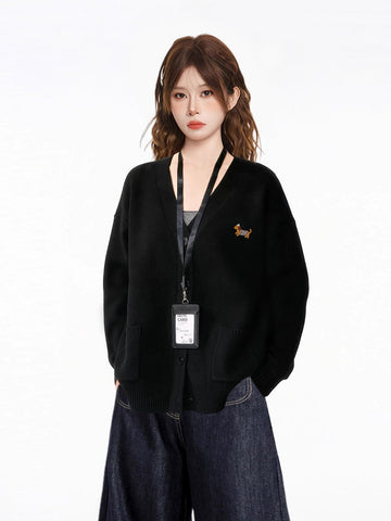 Woman wearing a black oversized knit cardigan with a V-neck, button-down front, and dual front pockets. The cardigan features an embroidered dachshund graphic on the left chest.