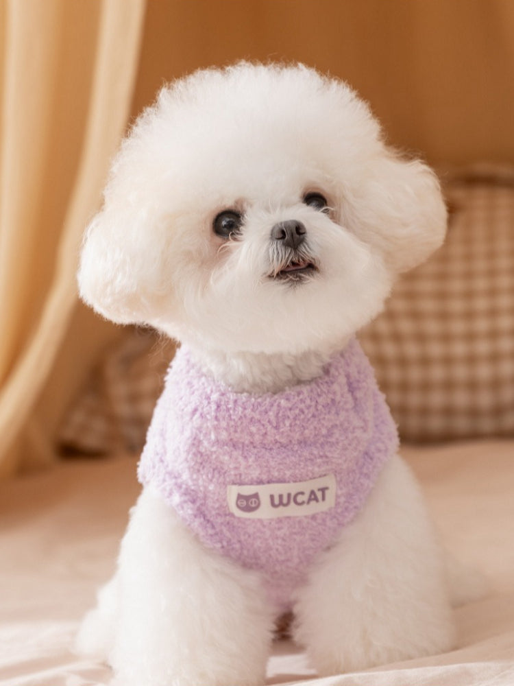 White bichon frise dog wearing a lavender fluffy dog sweater with a white "Wcat" logo patch on the chest.