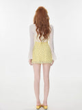 Rear view of a woman in a yellow floral print mini dress and white shrug, highlighting the back of the dress and the sheer sleeves of the shrug.