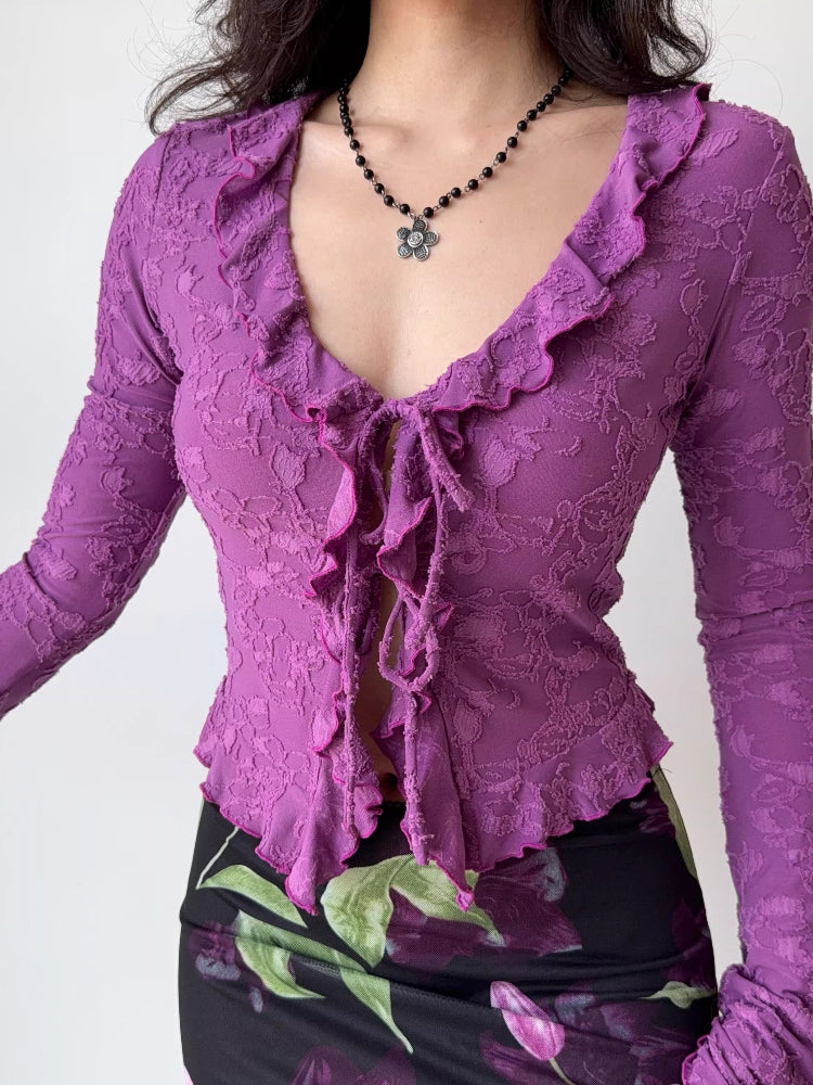 Front view of a woman in a purple textured mesh long-sleeve top with ruffled trim and a tie-front detail.