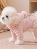 A Bichon Frise dog walking outdoors wearing a pink gingham hooded dog suit with a pink pom-pom accent and a leash.
