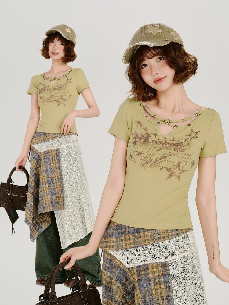 Full-body view of a woman wearing the light green graphic print t-shirt and a layered plaid and textured skirt, accessorized with a cap and a handbag.