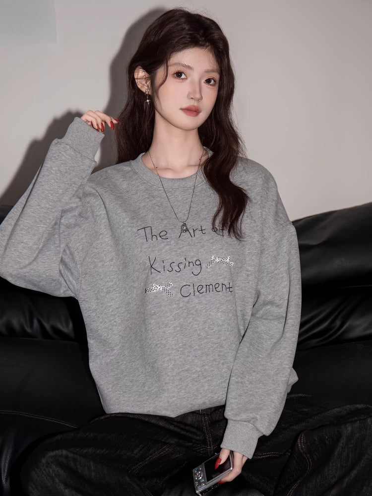 Woman wearing a grey long-sleeve sweatshirt with "The Art of Kissing Clement" graphic print.