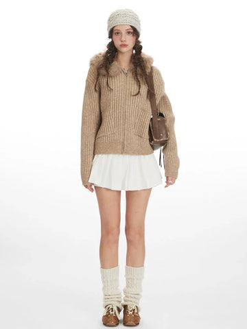Woman wearing a beige faux fur hooded cardigan jacket with a white pleated skirt