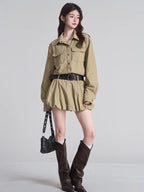Full body shot of a woman in a khaki camp collar shirt dress with a gathered skirt, accessorized with a black studded belt and dark brown boots.