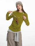 Model adjusting a long-sleeve olive green top with ruched side detail, grey layered vest, taupe wide-leg pants, yellow handbag, and striped headband.