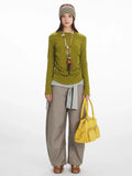 Model wearing a long-sleeve olive green top with ruched side detail, grey layered vest, taupe wide-leg pants, and a yellow handbag.