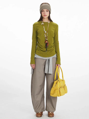 Model wearing a long-sleeve olive green top with ruched side detail, grey layered vest, taupe wide-leg pants, and a yellow handbag.