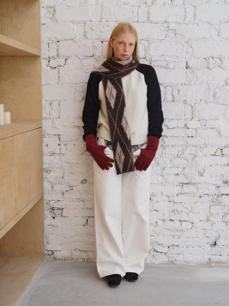 Full body shot of a model styled in a cream and black raglan sleeve sweater, argyle scarf, and white trousers.