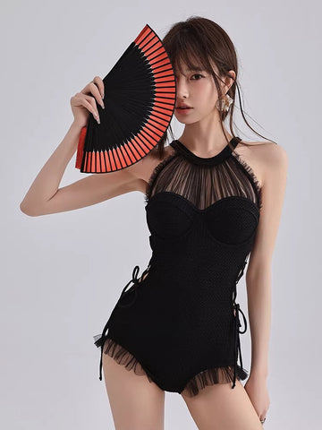 Woman wearing a black one-piece swimsuit with a sheer mesh halter neckline and ruffled hem, holding a red and black fan.