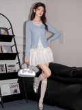 Woman standing next to a bookshelf and a sofa, wearing a light blue knit cardigan, white tiered skirt, and silver shoes.