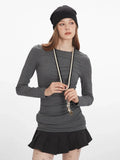Woman in a grey long-sleeved ruched top, black pleated mini skirt, patterned tights, black boots, and beanie, adjusting her necklace.