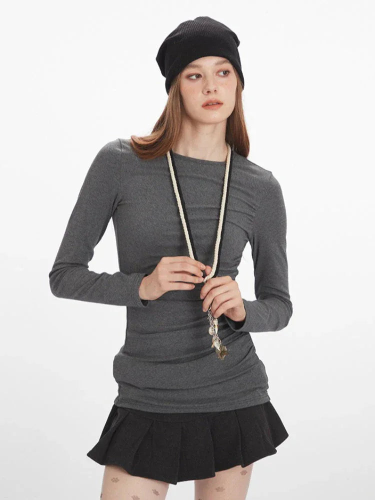Woman in a grey long-sleeved ruched top, black pleated mini skirt, patterned tights, black boots, and beanie, adjusting her necklace.
