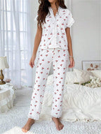 Woman wearing a white short-sleeve pajama top with a cherry print and lace trim, paired with white ribbed pajama pants featuring a cherry print and lace cuffs, standing in a bedroom.