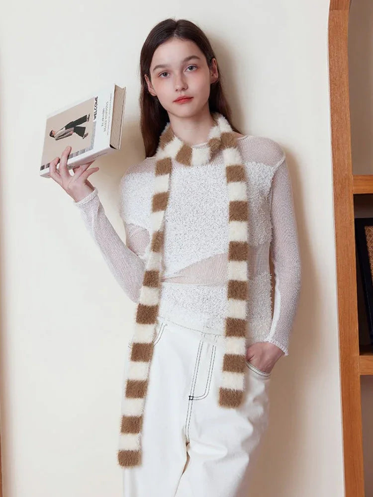 Woman holding a book and wearing a white long-sleeved top and white pants, with a brown and white striped fuzzy scarf.