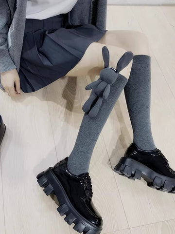 Model wearing grey knee-high socks with a 3D bunny charm on one leg, paired with a pleated skirt and platform shoes.