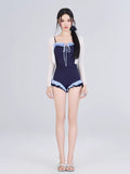 Woman wearing a navy blue one-piece swimsuit with light blue ruffled trim and a white sheer cardigan, standing