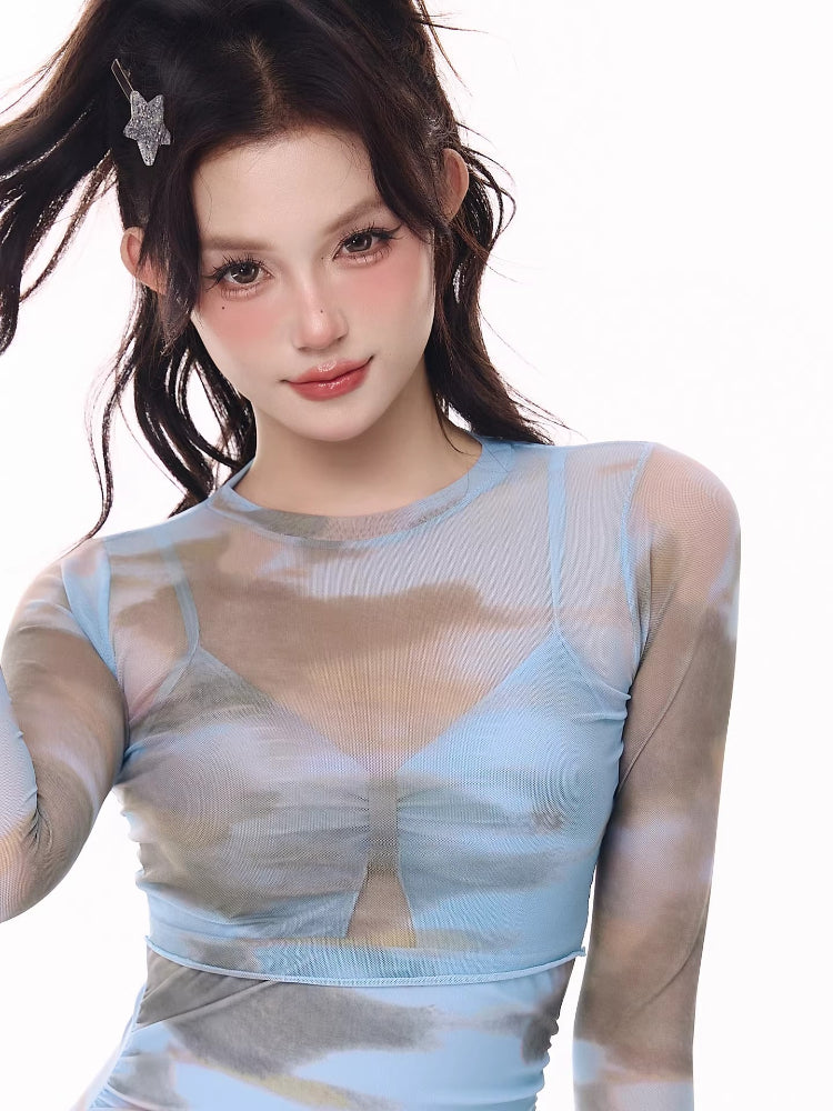 Woman wearing a blue and grey cloud print one-piece swimsuit with a sheer long-sleeve overlay and ruffled hem, posing with her hand in her hair.