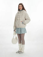 Full body shot of a woman in a cream fuzzy hooded jacket and a light blue pleated skirt.