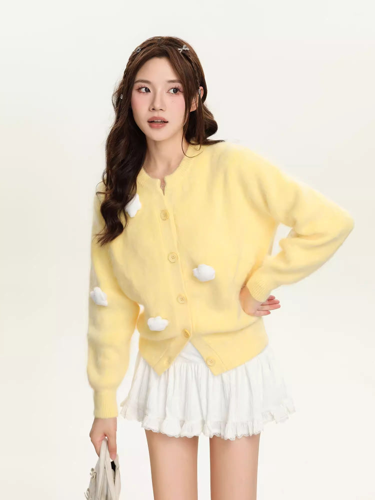 Full body shot of a woman in a yellow fuzzy knit cropped cardigan and white skirt, holding a handbag.